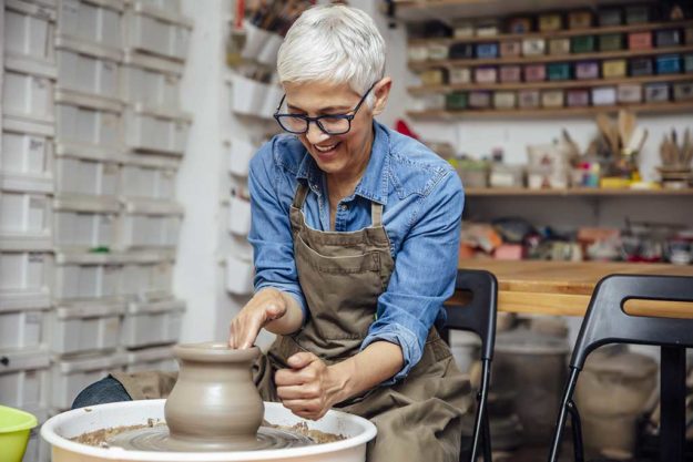 Senior female potter working on pottery wheel while sitting  in Senior female potter working on pottery wheel while sitting in