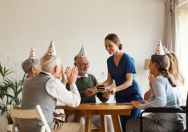 Young nurse bringing birthday cake with candle for senior man celebrating with aged friends sitting at table in nursing home. Happy elderly people in party hats clapping hands