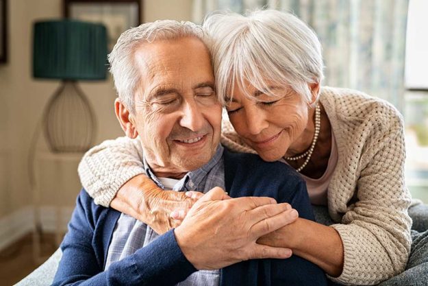 Romantic senior woman embracing from behind her husband sitting on sofa. Cute old couple in love hugging with closed eyes. Retired woman consoling her husband at home.