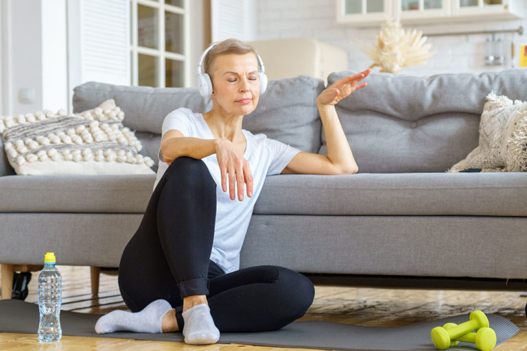 Old female relaxing after fitness activity at home, listening music. Stress relief activities Senior woman with closed eyes in headphones listen music in living room.