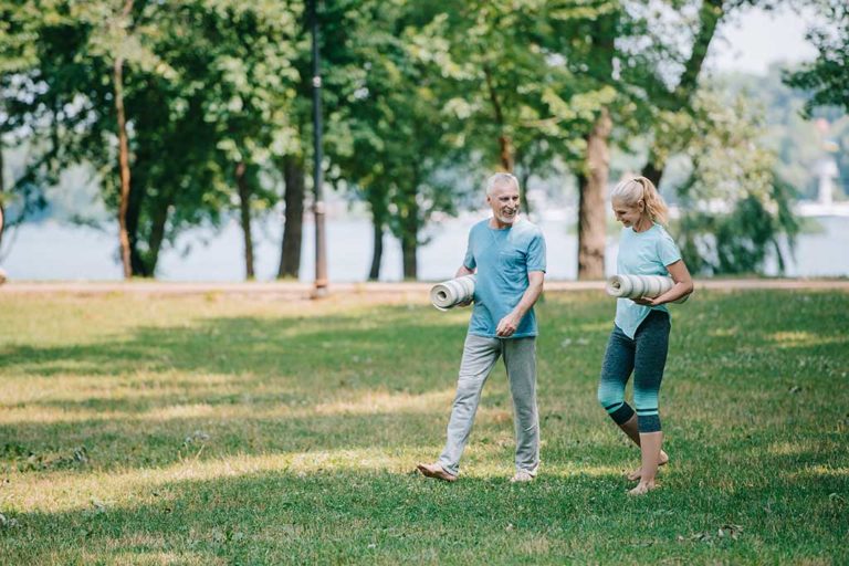 smiling mature man and holding yoga mats while walking in park together smiling mature man and holding yoga mats while walking in park together