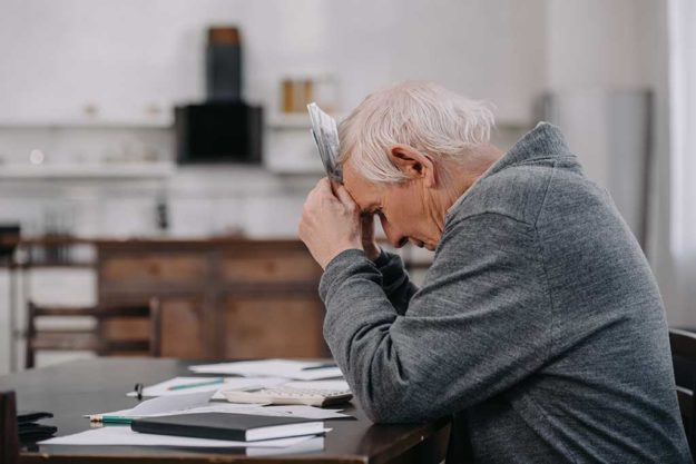 stressed senior man in casual clothes sitting at table with pape stressed senior man in casual clothes sitting at table with paperwork and holding money at home