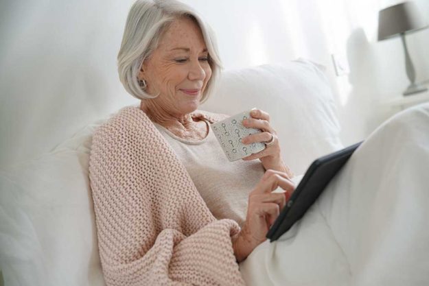Attractive senior woman relaxing in bed with hot drink and tabl Attractive senior woman relaxing in bed with hot drink and tablet