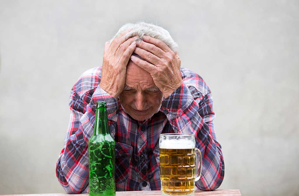 Senior drunk man with hangover Senior man with hangover holding his head at table with beer bottle and mug in front of him