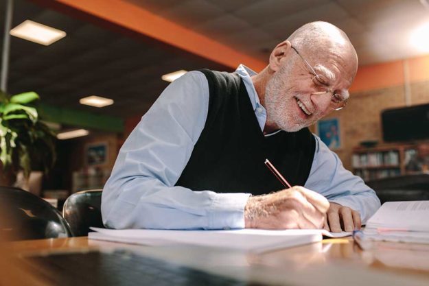 Senior man making notes in classroom Smiling senior man sitting in a library writing in his book. Cheerful elderly man learning sitting in a university classroom.