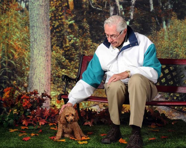 A senior man on a park bench in the fall petting his pet poodle.