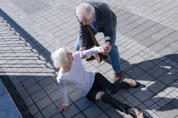 Pleasant woman tumbling over outdoors Do not afraid of falling down. Funny white-haired slim lady catching foot and receipting of relief from aged man who smiling
