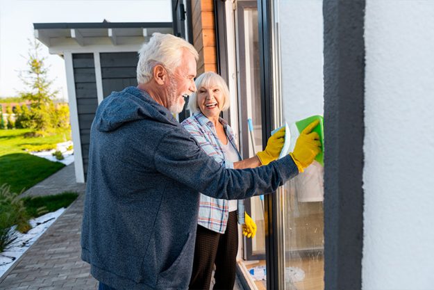 Cleaning together. Modern retired couple smiling broadly while washing windows outside together