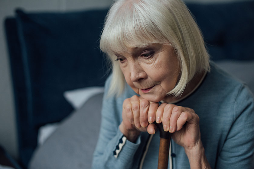 upset senior woman with hands on walking stick sitting on bed at home