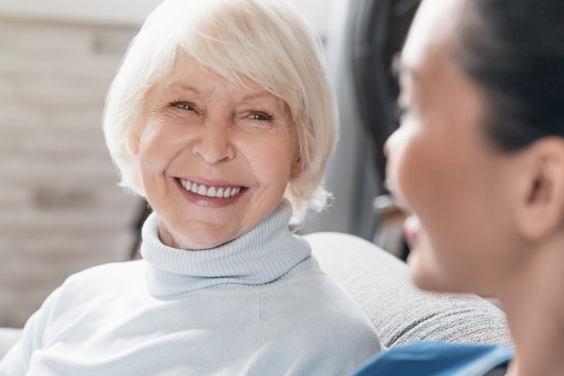 Close up of caregiver smiling and talking to aged woman while taking care of her at home