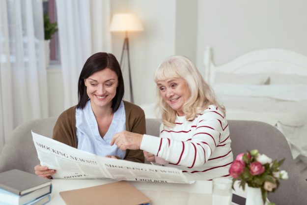 Inspired elder woman and caregiver examining newspaper