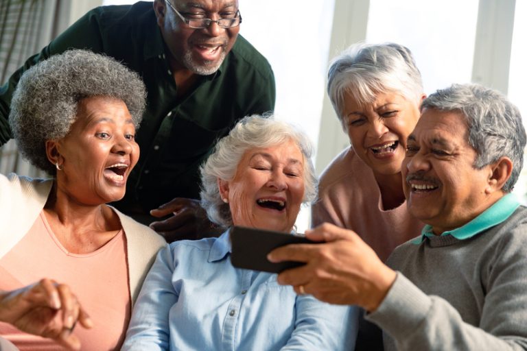 Five happy diverse senior friends sitting on sofa and looking at 6 Benefits Of Aging With Friends In Your Senior Years