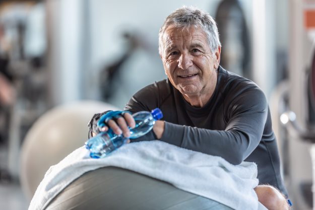 Elderly man resting on an exercise ball in the gym holds a water bottle looking at the camera. healthy-senior-lifestyle-habits-to-maintain-after-55-img