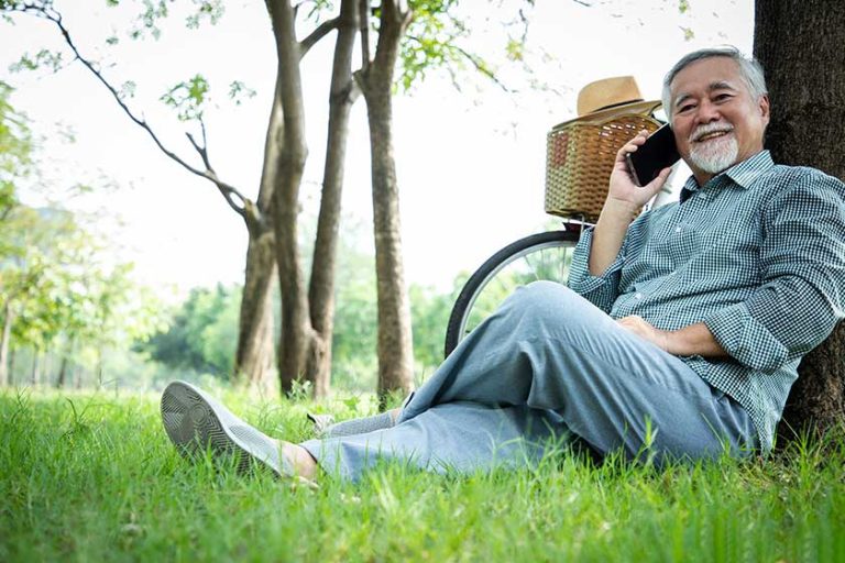 An elderly man sits under a tree talking on a cell phone with a bright