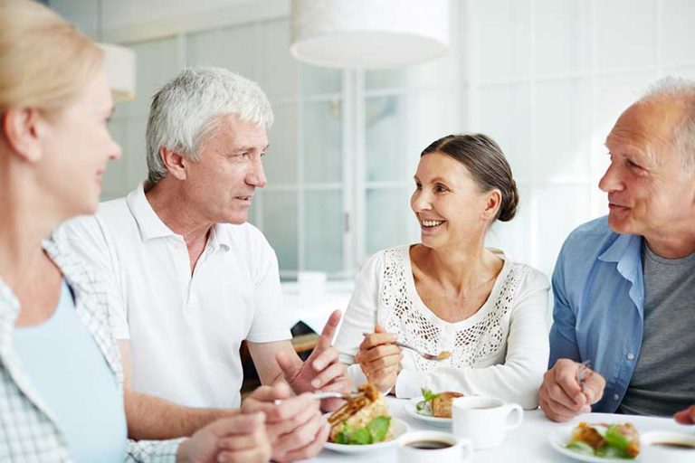 Friendly senior people gathered by table having talk by dessert with tea