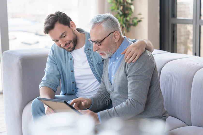 Caring loving adult son hugging embracing his old elderly senior father while he is showing his family photograph photo at home, telling stories of his youth. Happy father`s day! I love you, dad! Caring loving adult son hugging embracing his old elderly senior father while he is showing his family photograph photo at home, telling stories of his youth