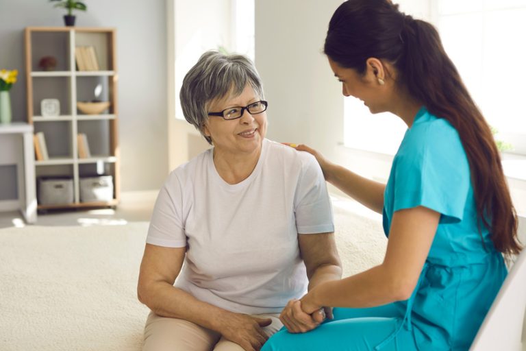 Friendly smiling nurse support old female patient sitting on home sofa