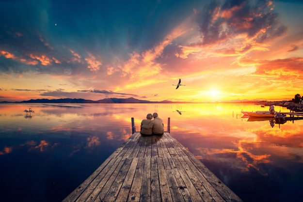 Senior couple seated on a wooden jetty, looking a colorful sunse Senior couple seated on a wooden jetty, looking a colorful sunse