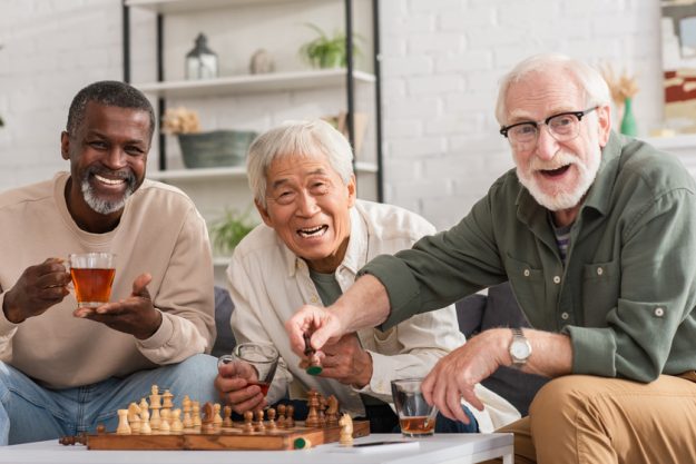 Cheerful multiethnic elderly friends looking at camera near chess and tea at home. Are You A First Time Resident In 55+ Communities? Here Are 4 Tips To Help You
