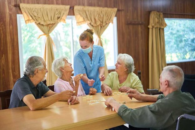 Caregiver wearing a face mask helps seniors play dominoes Caregiver wearing a face mask helps seniors play dominoes