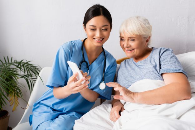cheerful asian nurse using mobile phone near happy senior woman