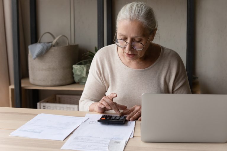 Older woman sitting at table using calculator manage personal budget What’s Included In Memory Care Costs?