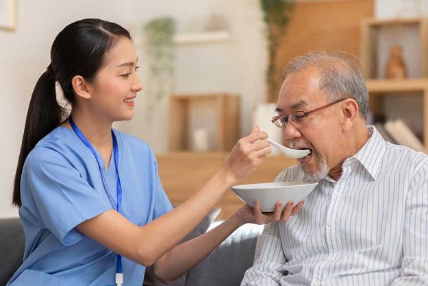 Young Asian nurse feeding breakfast to senior man at couch.
