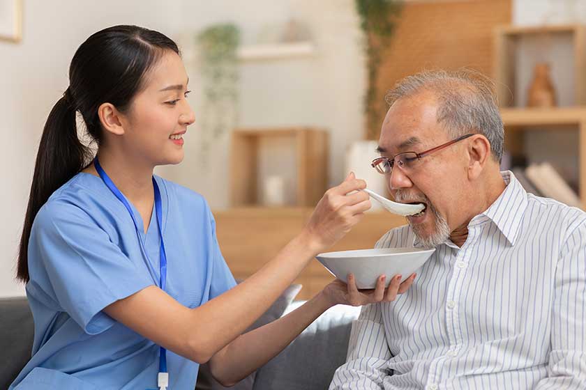 Young Asian nurse feeding breakfast to senior man at couch. Young Asian nurse feeding breakfast to senior man at couch.