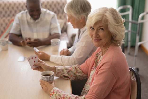 Front view of senior woman playing cards with senior people around table in living room at home How A Memory Care Facility Helps Your Loved Ones Overcome Gambling Addiction