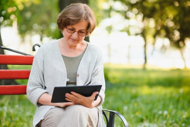 Attractive mature woman sitting on a bench holding and using a digital tablet in the park at summer day