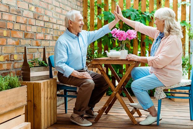 Cheerful senior couple giving high five while sitting near pink flowers on terrace Cheerful senior couple giving high five while sitting near pink flowers on terrace
