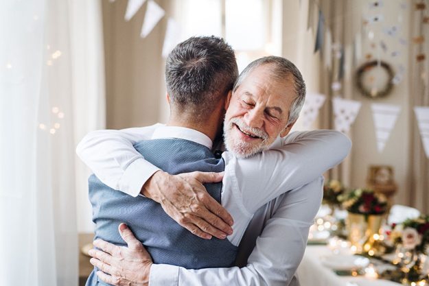 A senior and mature man standing indoors in a room set for a party, hugging