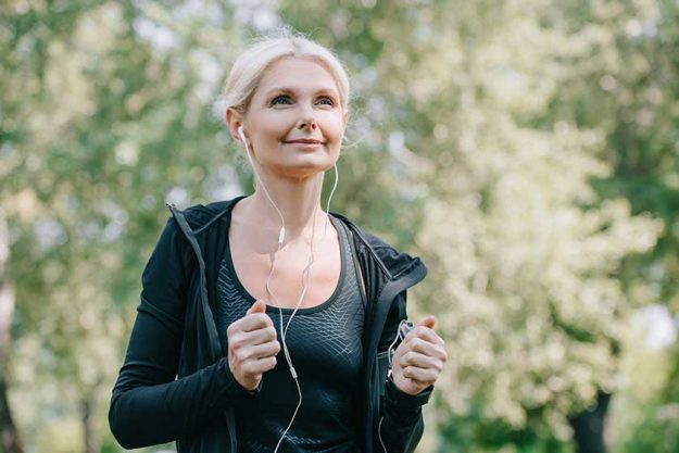 Beautiful mature sportswoman looking away while running in park and listening music in earphones