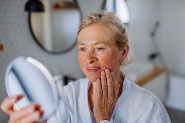 A beautiful senior woman in bathrobe looking at mirror and applying natural face cream in bathroom, skin care concept.