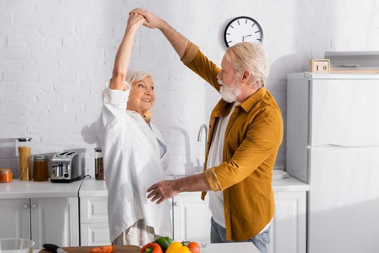 Cheerful senior couple dancing near fresh vegetables on kitchen table
