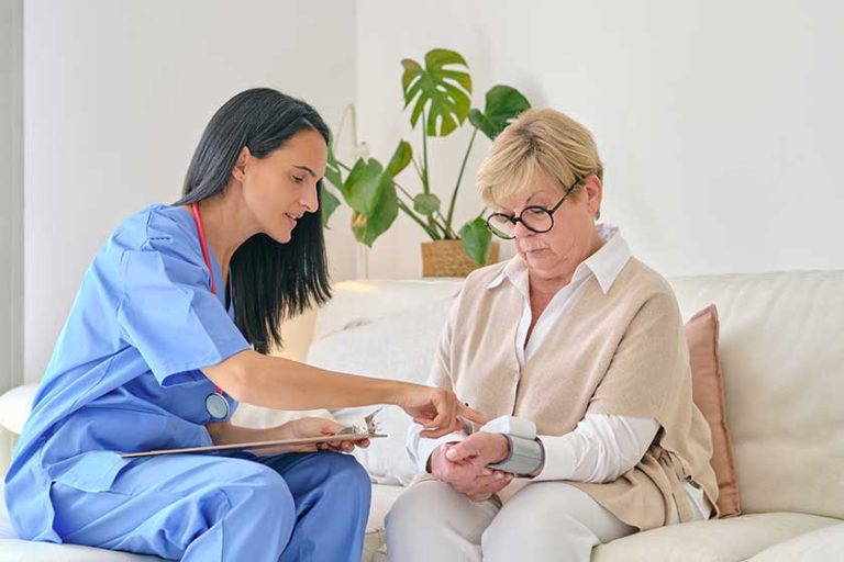 Female doctor in uniform checking up pulse rate on digital device on wrist of elderly patient in living room Female doctor in uniform checking up pulse rate on digital device on wrist of elderly patient in living room