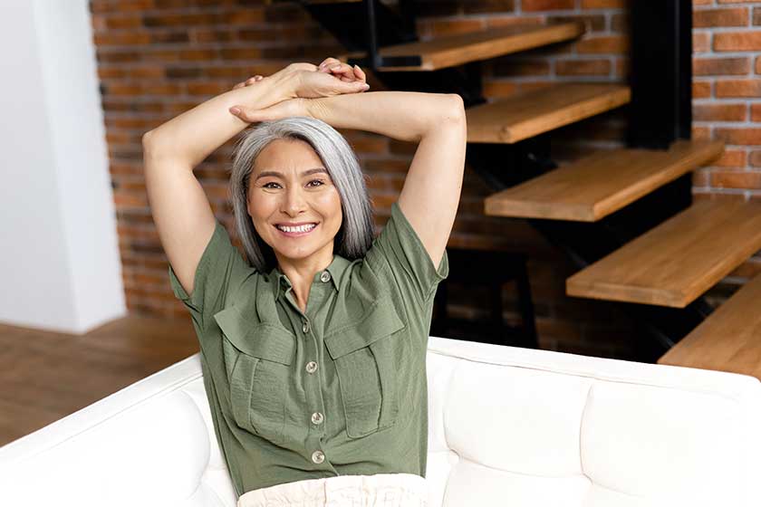 Positive middle-aged woman with long hair at the sofa in modern apartment. 50s asian female resting at home, looks at the camera Positive middle-aged woman with long hair at the sofa in modern apartment. 50s asian female resting at home, looks at the camera