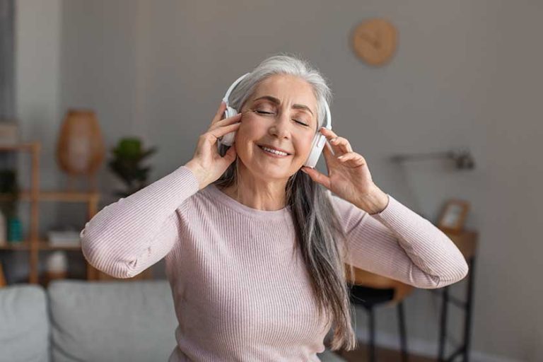 Smiling pretty european mature woman with gray hair in wireless headphones enjoys favorite music and free time in room interior. Audio app at comfort home alone and new device during covid-19 pandemic Smiling pretty european mature woman with gray hair in wireless headphones enjoys favorite music and free time in room interior. Audio app at comfort home alone and new device during covid-19 pandemic
