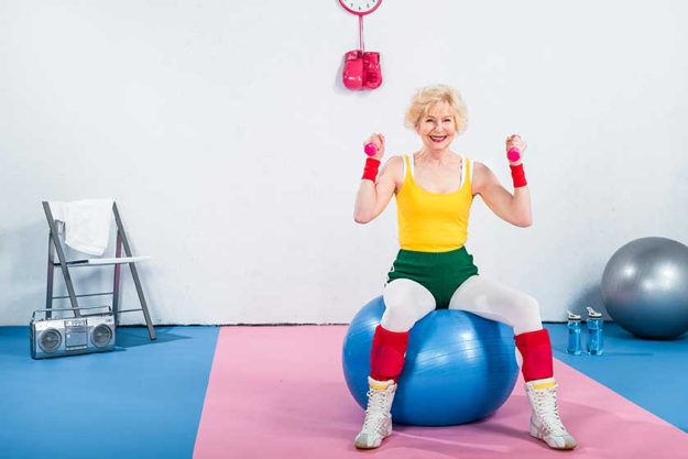 Smiling sportive senior woman sitting on fitness ball and exercising with dumbbells Smiling sportive senior woman sitting on fitness ball and exercising with dumbbells
