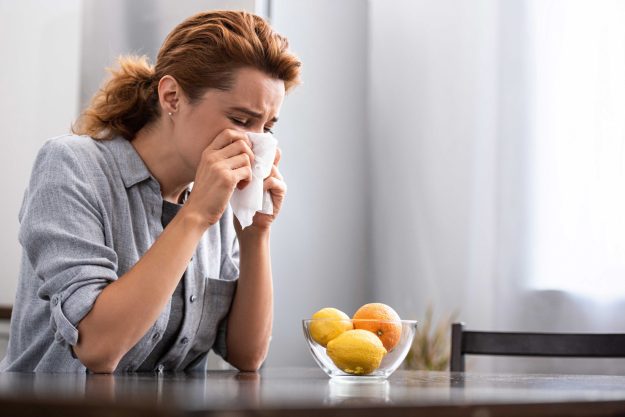 Woman with runny nose sneezing in tissue near orange and lemons in bowl Woman with runny nose sneezing in tissue near orange and lemons in bowl