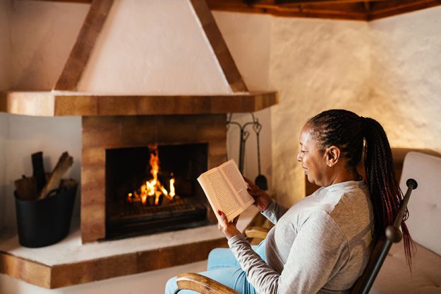 senior african woman reading book while warming front fireplace her