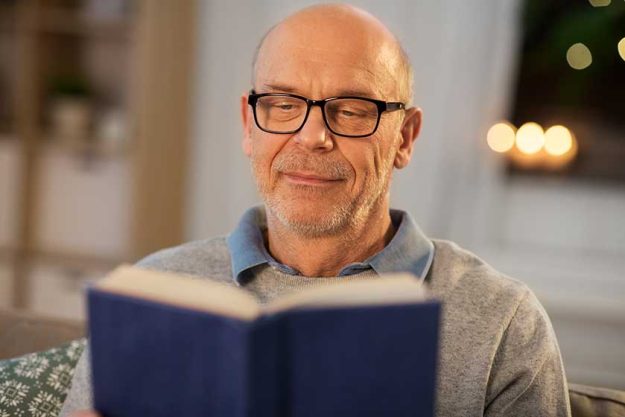 happy bald senior man on sofa reading book at home
