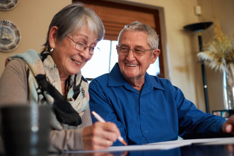 Smiling Senior Couple At Home Sitting At Table Checking Personal Finances