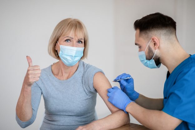 Mature woman in face mask approving of covid 19 vaccination, showing thumb up during coronavirus vaccine injection