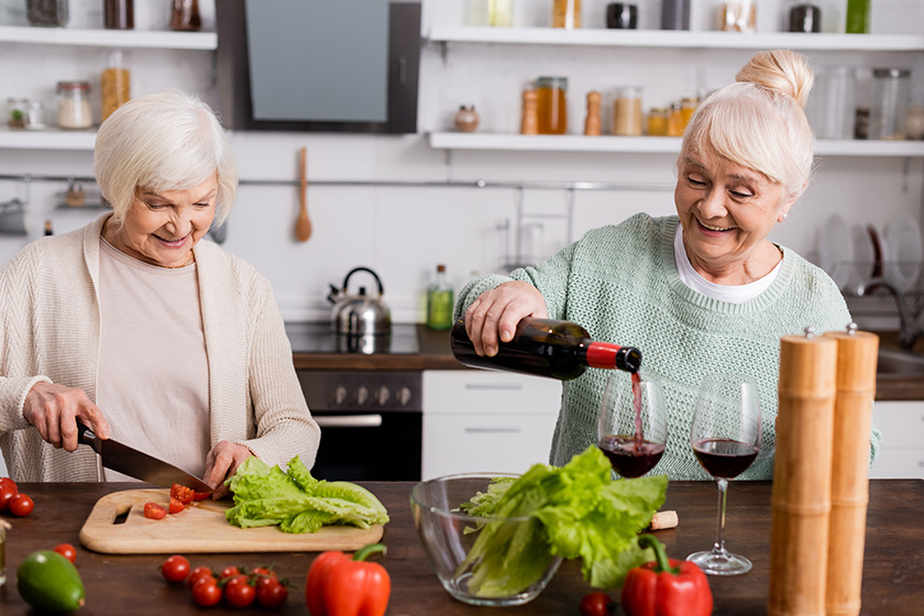 happy senior woman pouring red wine glass retired friend cutting
