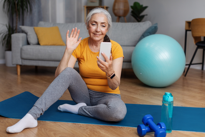 Senior woman with smartphone making video call with personal couch while sitting on fitness mat at home Senior woman with smartphone making video call with personal couch while sitting on fitness mat at home