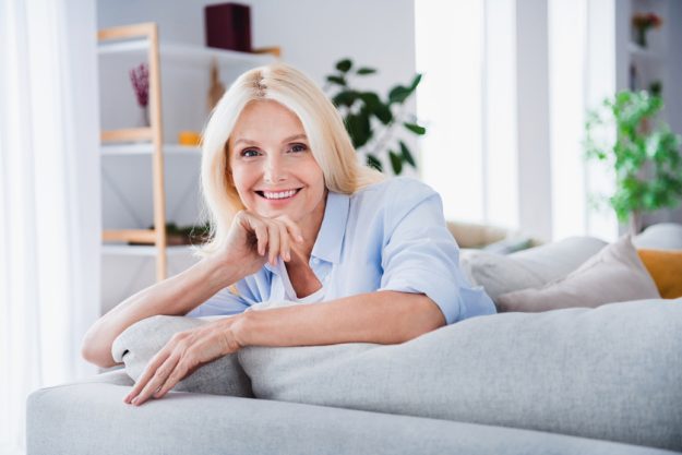 Photo portrait of charming retired woman sit sofa charming toothy smile enjoy weekend stylish home decor living room modern apartment