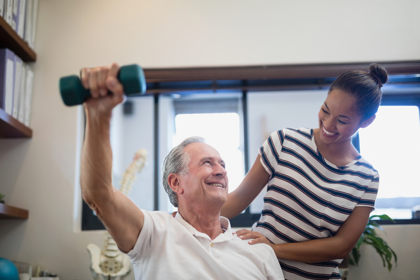 Smiling senior male patient lifting dumbbell while looking at female doctor Smiling senior male patient lifting dumbbell while looking at female doctor