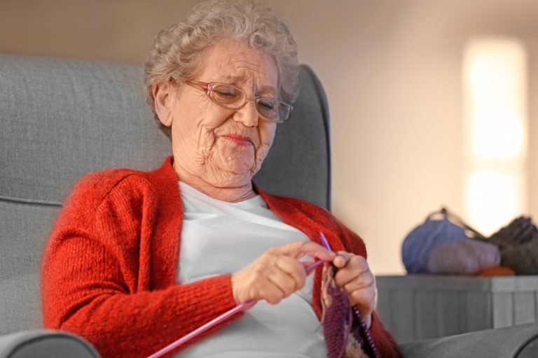 Senior woman knitting warm scarf in armchair at home