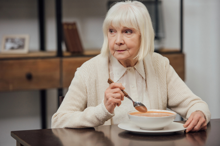 Lonely senior woman sitting at table and eating cream soup at home